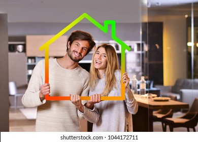 Portrait Of A Couple In Love Who Have Just Bought A House And Is Moving. The Bride And Groom Smile And Hold And Hold The Outline Of A House The In Their Hands. Concept Of: Home, Family, Ecology