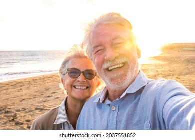 Portrait of couple of mature and old people enjoying summer at the beach looking to the camera taking a selfie together with the sunset at the background. Two active seniors traveling outdoors.
