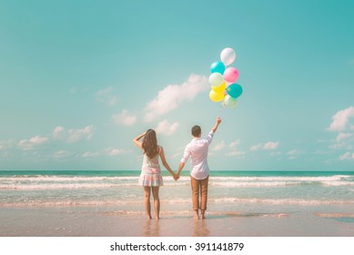 Portrait of couple of young happy married hipsters in trendy vintage clothes standing together on the beach with balloons. Sunny summer day. Pastel colors tone 