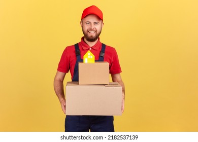 Portrait Of Courier Man In Blue Coveralls And Red T-shirt Holding Cardboard Boxes With Paper House On Top, Movement Company, Safe Delivery. Indoor Studio Shot Isolated On Yellow Background.