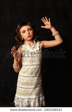 Portrait of cover little doll girl gesture hands posing at black background, frozen position. Adorable child 7-8 year old isolated in shadow, darkness. Theatre performance concept. Copy ad text space