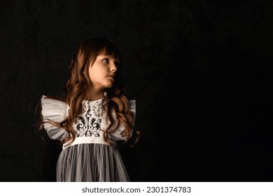Portrait of cover little girl in doll image isolated in shadow, frozen look looking away. Charming kid 9-10 year old with curly hair at black background. Fashion style concept. Copy ad text space