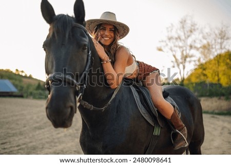 Portrait of cowgirl with stetson hat sitting on horse backs at homestead and smiling at the camera. Smiling rider in western styled outfit sitting on horse backs and looking at the camera at ranch.