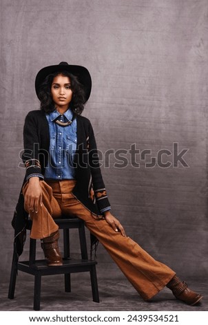 Portrait, cowgirl or woman in studio, wild west culture and cool fashion in clothing on grey background. Young female person, western lady and stylish model with pride, boho style and chair stool