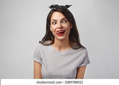 Portrait Of Crazy Beautiful Young Caucasian Female With Dark Hair Marking Goofy Face, Squinting Eyes And Sticking Out Tongue While Fooling Around, Posing In Studio. Human Facial Expressions