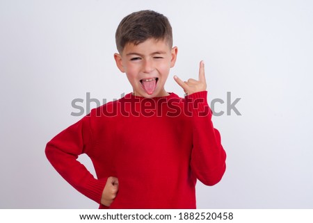Portrait of a crazy Cute Caucasian kid boy wearing knitted sweater against white wall showing tongue horns up gesture, expressing excitement of being on concert of band.