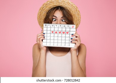 Portrait of a crazy funny girl in summer hat holding and hiding behind a periods calendar isolated over pink background
