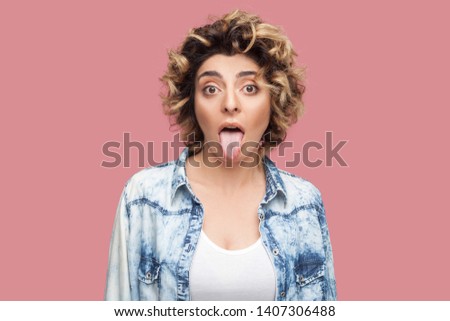 Portrait of crazy or shocked funny young woman with curly hairstyle in casual blue shirt standing, tongue out and looking at camera with big eyes. indoor studio shot, isolated on pink background.