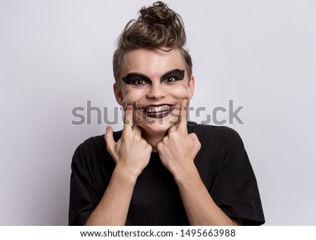 Portrait of crazy teen boy with spooking make-up on grey background. Young teenager in style of punk goth dressed in black looking at camera with big smile. Problems of transitional age.