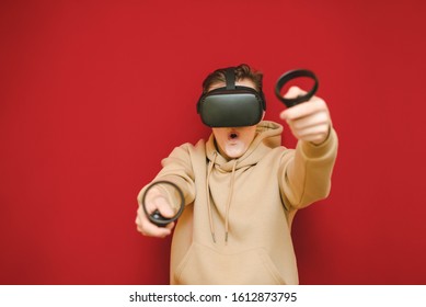 Portrait Crazy Young Man In VR Helmet And Controllers In Hands Playing Video Games On Red Background.Mad Gamer Plays VR Games On A Virtual Reality Helmet And Looks At The Camera With A Surprised Face