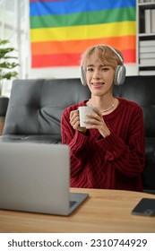 Portrait of a creative and confident young Asian gay man freelancer is working from home, holding a coffee mug and listening to music through headphones in the living room. - Shutterstock ID 2310744929