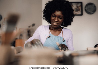 Portrait of a creative craftswoman sitting in modern studio on pottery class and learning pottery and clay work. An african american smiling pottery course attendee modeling clay on pottery course. - Powered by Shutterstock