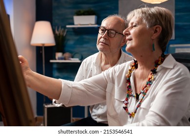 Portrait Of Creative Elderly Couple Drawing In Pencil Sitting Down In Front Of Easel Talking About Composition In Home Art Studio. Older Man Looking At Gray Haired Woman Doing Charcoal Sketch.