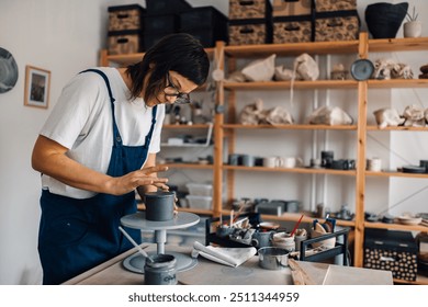 Portrait of creative female pottery designer in overalls standing at pottery workshop and making clay mug. Precise female artisan standing at pottery workshop and making earthenware. Copy space. - Powered by Shutterstock