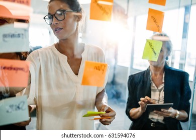 Portrait Of Creative Professionals Looking Over A Post It Note Wall And Discussing. Female Executives Standing At The Office Behind Glass Wall With Sticky Notes.