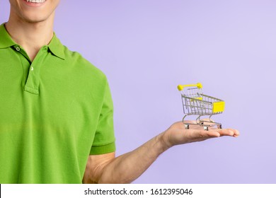 Portrait Of Cropped Young Man In Green T-shirt Holding Small Toy Truck For Shopping From Hypermarket, Isolated Over Purple Background