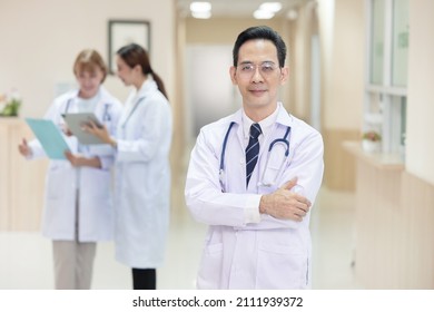 Portrait Of Crossed Arm Asian Doctor In A Gown Standing In A Hospital With Two Female Doctors  In The Background