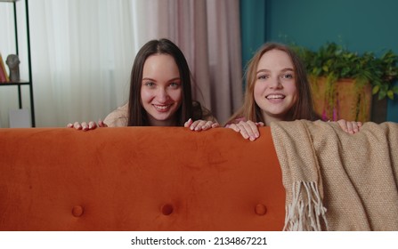 Portrait Of Cunning Girls Friends Siblings Playing Hide And Seek Peekaboo Game Near Sofa Looking At Camera And Smiling In Positive Mood. Two Female Women Couple Family Trying To Scare, Prank At Home