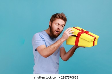 Portrait of cunning handsome bearded man unboxing yellow gift box, looking inside with curious facial expression, birthday present. Indoor studio shot isolated on blue background. - Shutterstock ID 2187570953
