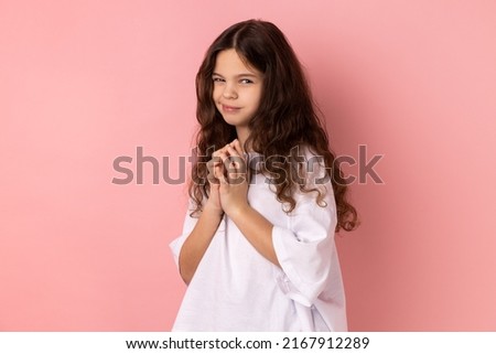 Portrait of cunning little girl in T- shirt having tricky plans, looking at camera with cheating face, smirking mysteriously, scheming cheats. Indoor studio shot isolated on pink background.