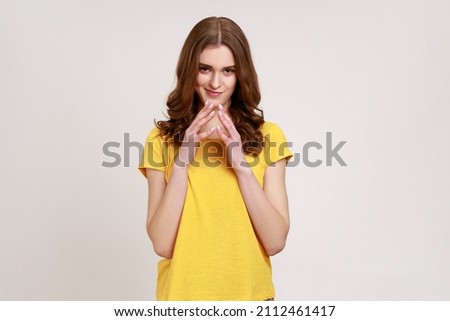 Portrait of cunning smart cute teenager girl with brown hair in yellow casual T- shirt standing and looking at camera with sly smile, having tricky plan. Indoor studio shot isolated on gray background