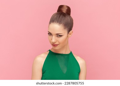 Portrait of cunning woman with bun hairstyle looking at camera with sly facial expression, having devil plant or preparing prank, wearing green dress. Indoor studio shot isolated on pink background.