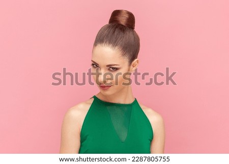 Portrait of cunning woman with bun hairstyle looking at camera with sly facial expression, having devil plant or preparing prank, wearing green dress. Indoor studio shot isolated on pink background.