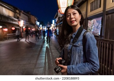 Portrait Of Curious Asian Female Tourist Carrying Camera And Looking Into Distance With Smile While Exploring Geisha District On The Busy Hanamikoji Street In Gion Kyoto Japan At Night