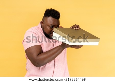 Portrait of curious attentive man wearing pink shirt holding present box in hands and looking inside with serious expression and big eyes. Indoor studio shot isolated on yellow background.