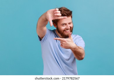 Portrait of curious bearded man gesturing picture frame with hands, looking through fingers and focusing on interesting moment, taking photo. Indoor studio shot isolated on blue background.
