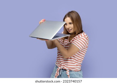 Portrait of curious blond woman wearing striped T-shirt holding half opened laptop, spying , finding important or forbidden information. Indoor studio shot isolated on purple background.
