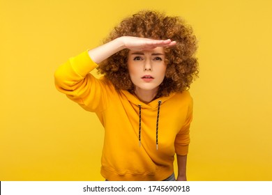Portrait of curious curly-haired woman in urban style hoodie holding hand above eyes and peering into distance, looking far away, expecting and searching someone on horizon. studio shot isolated - Shutterstock ID 1745690918