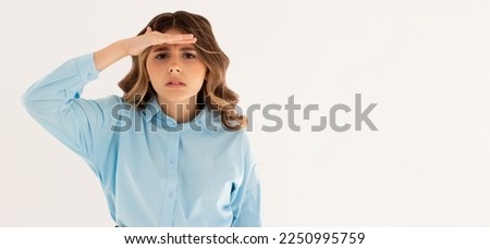 Portrait of curious wavy-haired woman in blue shirt holding hand above eyes and peering into distance, looking far away, expecting and searching someone on horizon. studio shot isolated