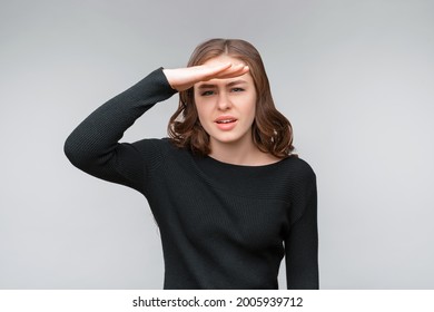 Portrait of a curious young brunette woman looking far away with hand at her forehead, trying to see something far away. Studio shot, gray background. Human emotions, facial expression concept