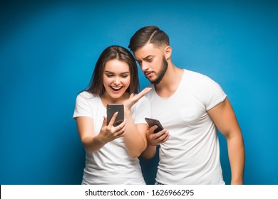 Portrait of curious young couple man and woman in basic t-shirts smiling while peeking at each other cellphones isolated over background.