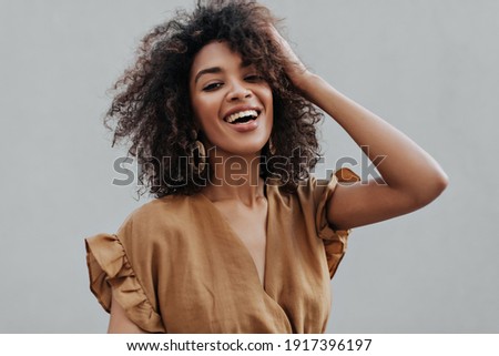 Portrait of curly brunette African dark-skinned woman in beige top smiling and ruffling hair on isolated grey background.