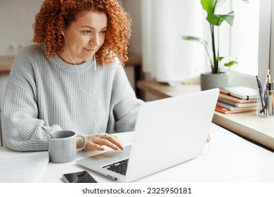 Portrait of curly haired redhead caucasian female in wired headphones spending leisure time using laptop during coffee break, sitting at kitchen table with smartphone, visiting popular web pages - Shutterstock ID 2325950181