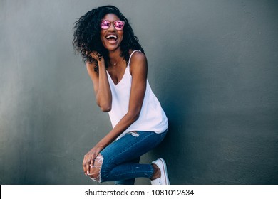 Portrait of a curly haired woman wearing fashionable clothes standing against a wall. Happy looking woman standing against a wall with arms crossed wearing sunglasses.