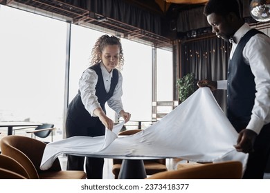 Portrait of curly haired young woman as classic server preparing restaurant for opening and folding tablecloths, copy space - Powered by Shutterstock