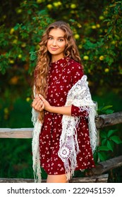 Portrait of a curly long hair Beautiful girl in a red velvet dress and shawl posing in apple garden. Art work of romantic woman .Pretty tenderness model looking at camera.