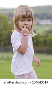 Portrait Of A Cut Child Girl With A Finger In Her Mouth Biting Her Nail Standing In Park Or Garden In Summer.