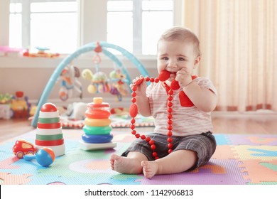 Portrait Of Cute Adorable Blond Caucasian Smiling Child Boy With Blue Eyes Sitting On Floor In Kids Children Room. Little Baby Playing With Teething Toys Jewelry. Early Education Development Concept.