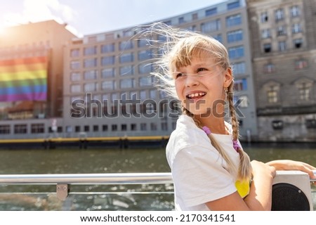 Portrait of cute adorable blond child girl stand on board river Spree Cruise trip boat with LGBTQ flag poster on building on background on brigh sunny day Berlin . Pride day fest celebraion Germany