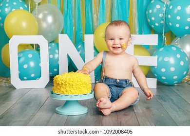 Portrait of cute adorable Caucasian baby boy in jeans pants celebrating his first birthday. Cake smash concept. Child kid sitting on floor in studio eating tasty yellow dessert 