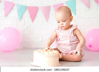 Portrait of cute adorable caucasian baby girl in pink jumpsuit celebrating her first birthday with cake and balloons, first year cake destruction concept. The girl is eating her birthday cake.