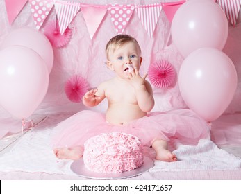 Portrait of cute adorable Caucasian baby girl with blue eyes in pink tutu skirt celebrating her first birthday with gourmet cake and balloons looking in camera, cake smash first year concept