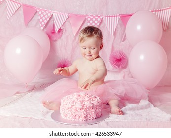 Portrait of cute adorable Caucasian baby girl with blue eyes in pink tutu skirt celebrating her first birthday with gourmet cake and balloons looking away, cake smash first year concept