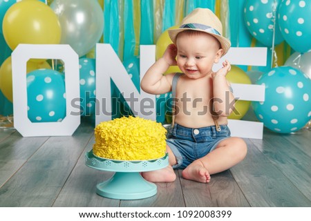 Portrait of cute adorable Caucasian baby boy wearing jeans pants and hat celebrating his first birthday. Cake smash concept. Child kid sitting on floor in studio eating tasty yellow dessert 