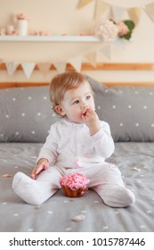 Portrait of cute adorable Caucasian blonde baby girl in white onesie celebrating her first birthday eating pink cupcake. Home indoors cake smash first year concept