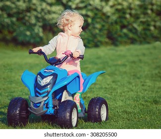 Portrait Of Cute Adorable Happy Little Blond Caucasian Boy Girl Child Driving Blue Electric Car In Park Outside On Sunny Windy Summer Day, Toned With Instagram Filters